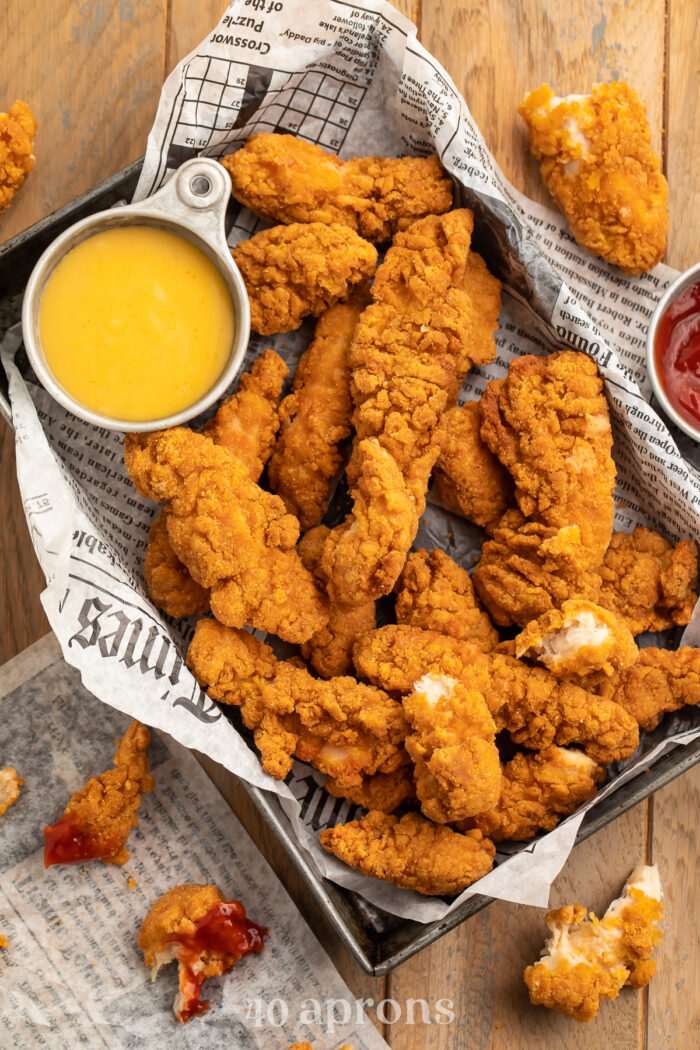Overhead view of a pan lined with newspaper filled with chicken tenders and a small bowl of yellow honey mustard dipping sauce.