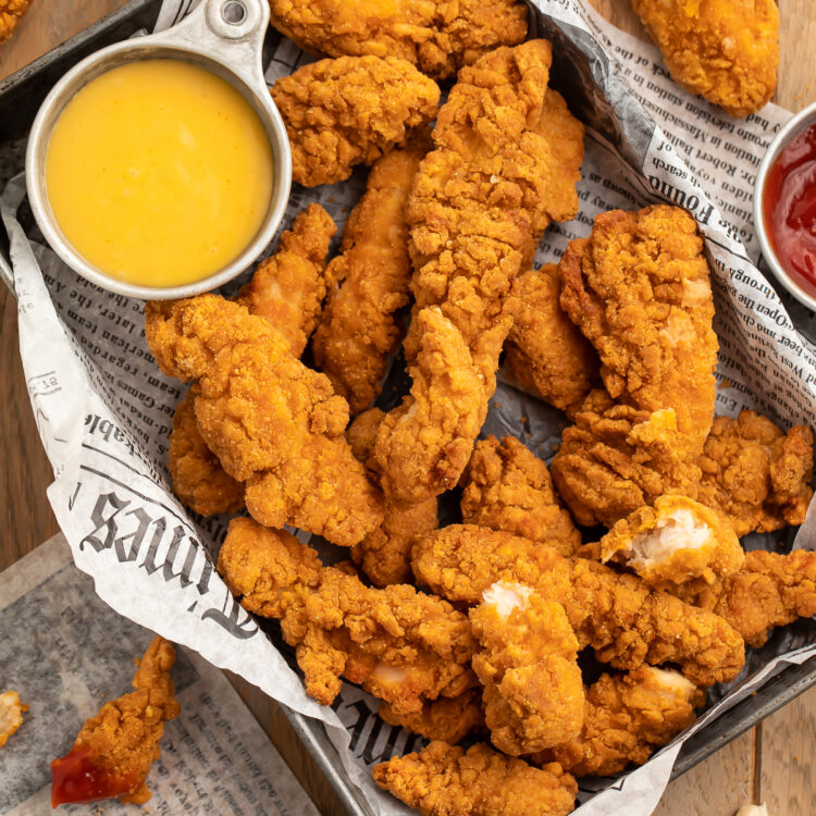 Overhead view of a pan lined with newspaper filled with chicken tenders and a small bowl of yellow honey mustard dipping sauce.