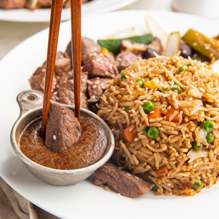 Bite-sized steak held by chopsticks being dipped into a ramekin of brown sauce next to a mound of veggie fried rice on a white plate.