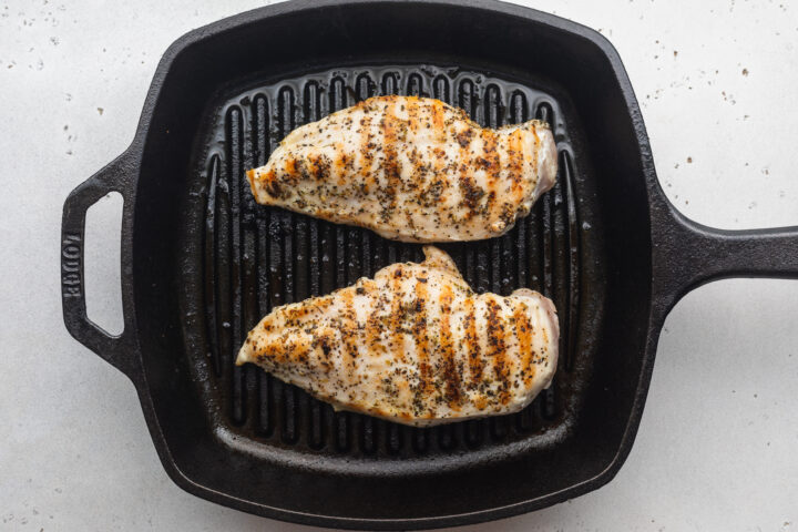 Overhead view of two grilled chicken cutlets in a grill pan on a neutral countertop.