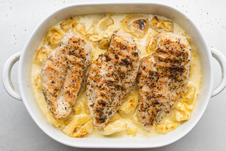 Overhead view of asiago tortelloni alfredo topped with 3 grilled chicken cutlets in a large oval baking dish.