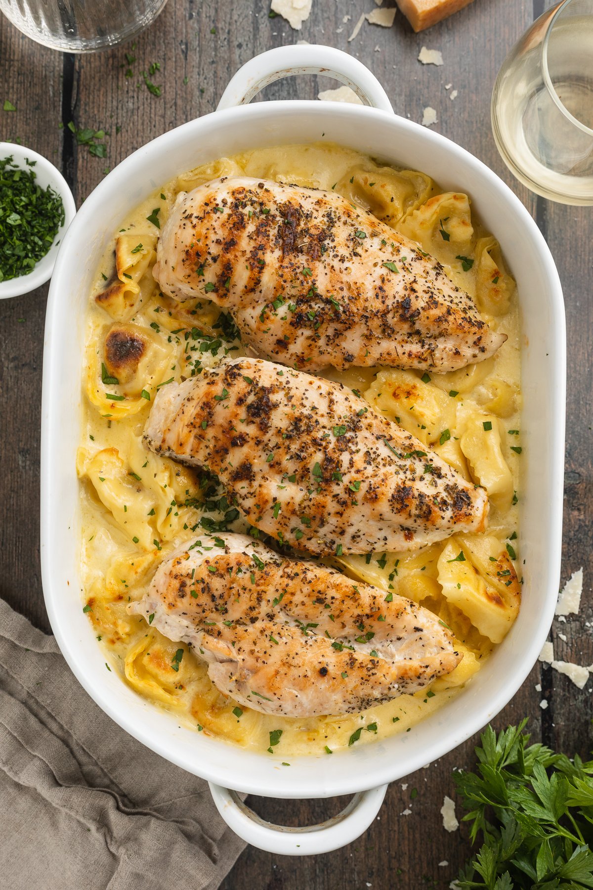 Overhead view of a large oval white baking dish holding copycat asiago tortelloni alfredo topped with 3 grilled chicken breasts.