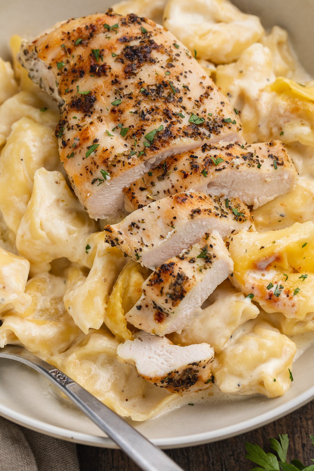 Overhead, close-up view of grilled chicken breast slices atop a bed of asiago tortelloni alfredo in a large bowl.