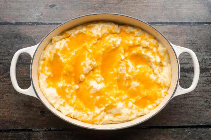 Overhead view of creamy, cheesy mashed potatoes topped with cheddar in a large oval baking dish with handles on each end.