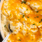 Overhead view of a large dish of cheesy mashed potatoes. A large silver spoon rests in the potatoes.