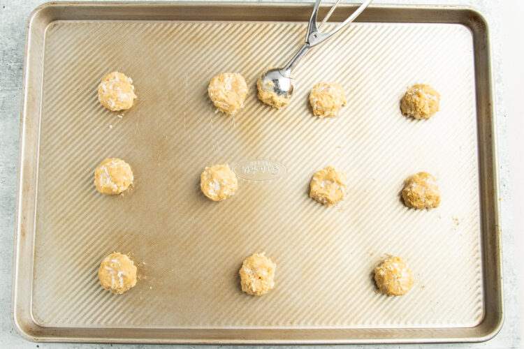 Baking sheet lined with parchment paper and covered with equally-sized scoops of cookie dough and a cookie scoop.