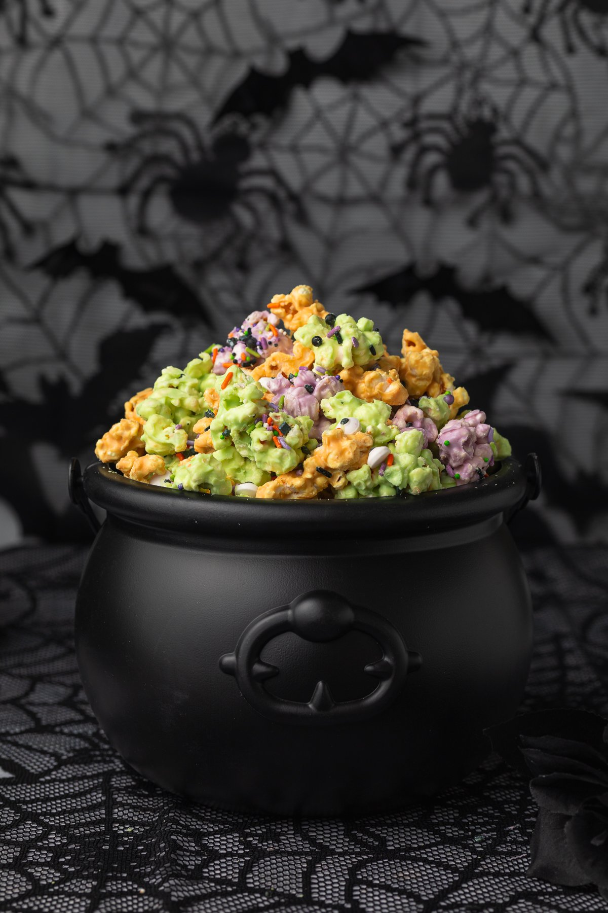 Side view of a plastic cauldron filled with orange, purple, and green white chocolate covered popcorn clusters with sprinkles and candy eyes.