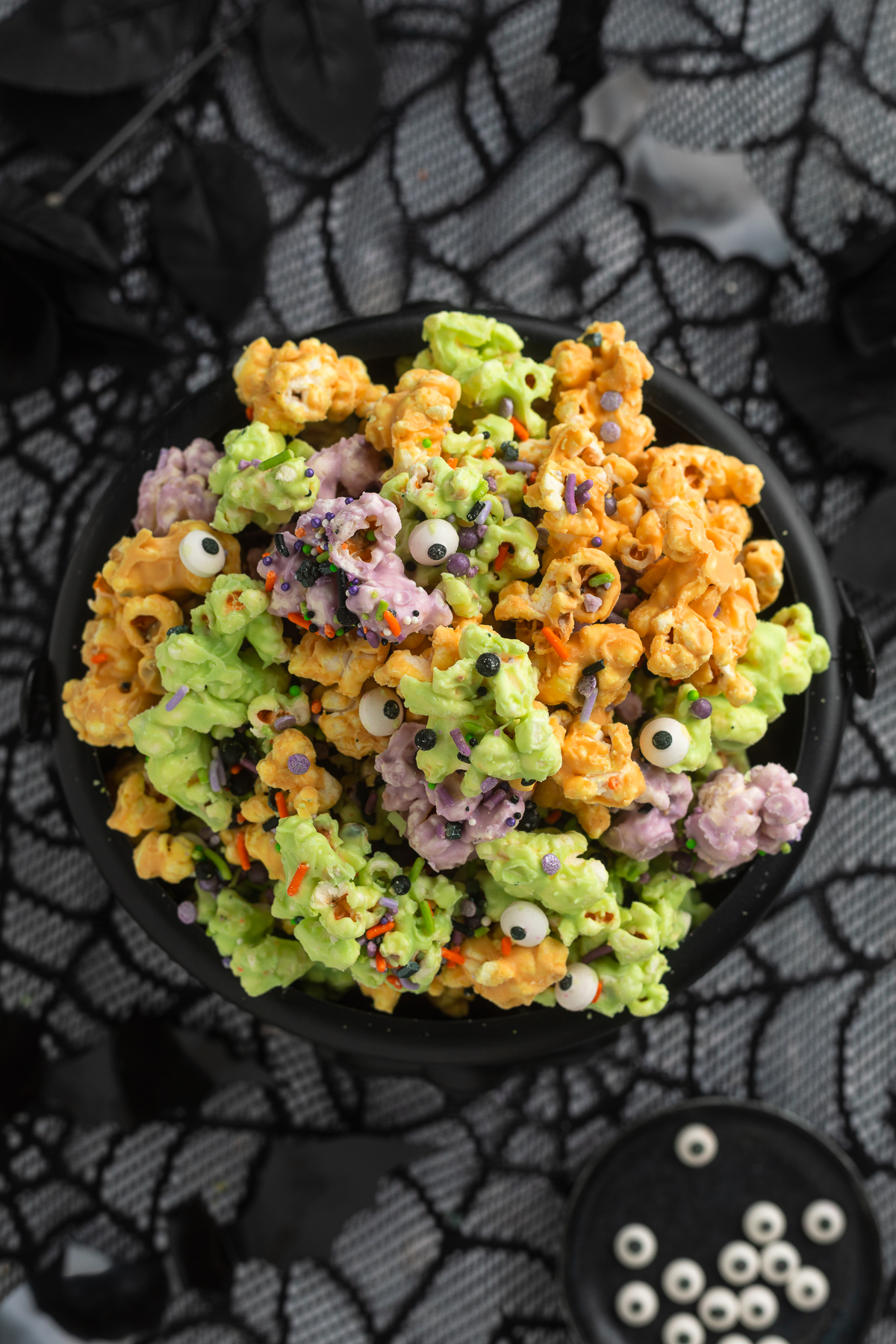 Overhead view of a plastic cauldron filled with orange, purple, and green white chocolate covered popcorn clusters with sprinkles and candy eyes.