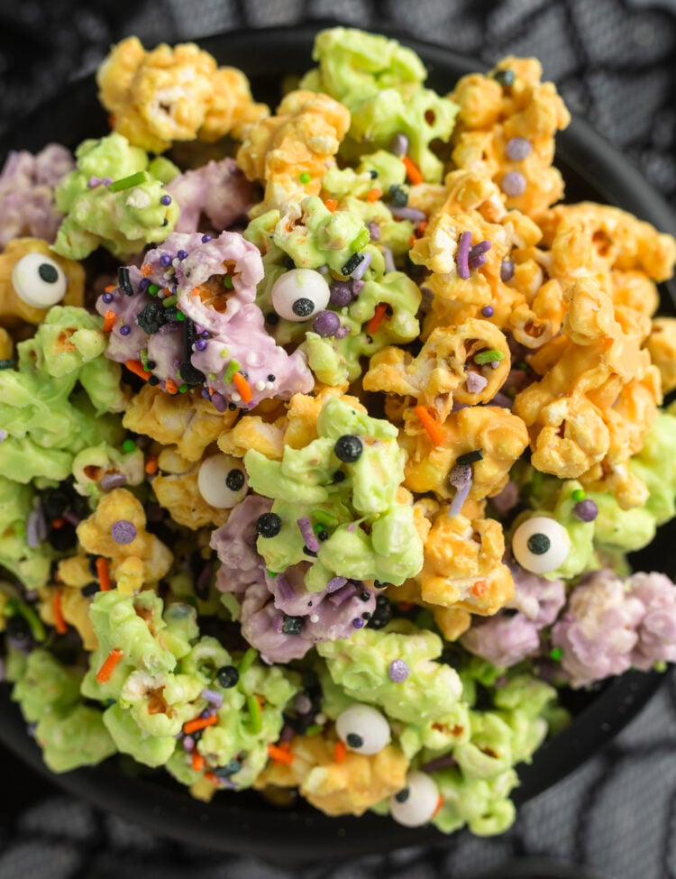Close-up, overhead view of a plastic cauldron filled with orange, purple, and green white chocolate covered popcorn clusters with sprinkles and candy eyes.
