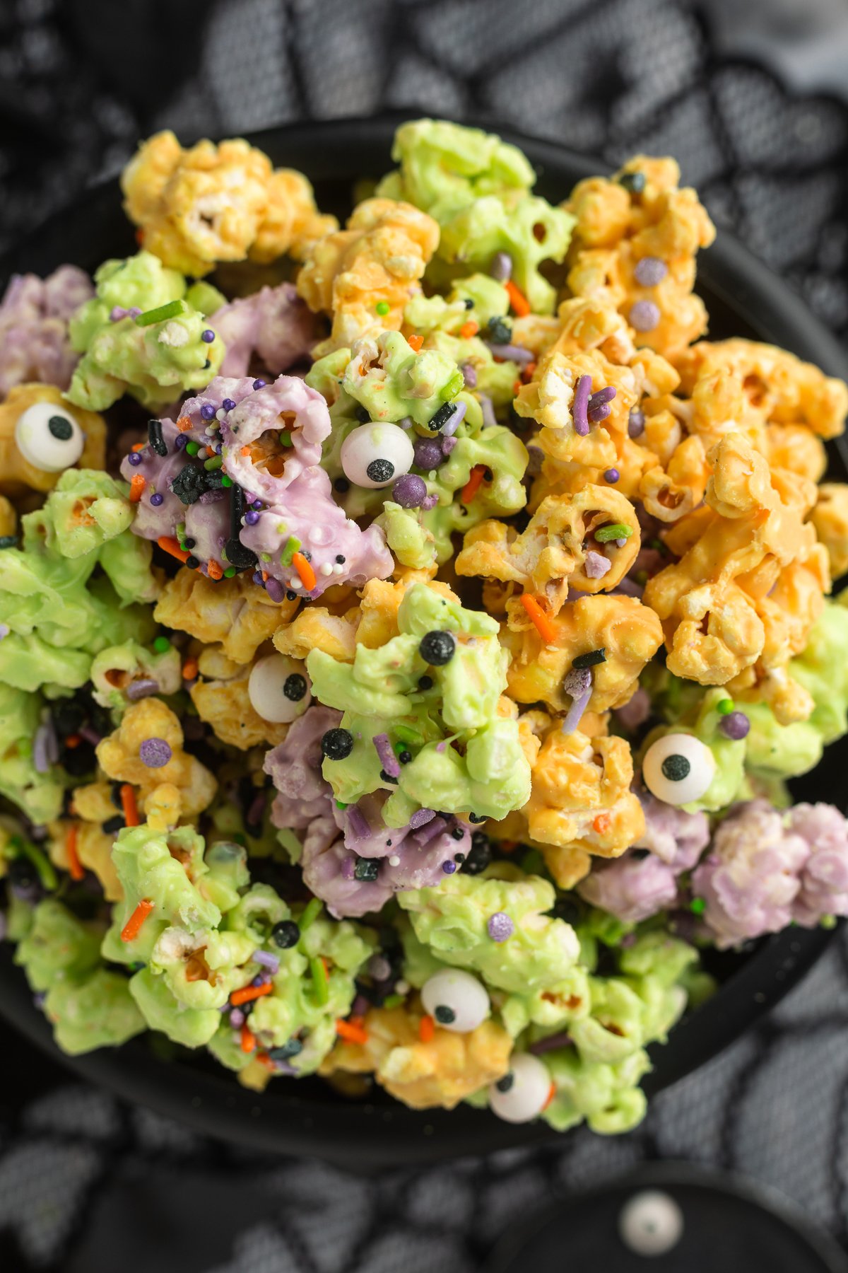 Close-up, overhead view of a plastic cauldron filled with orange, purple, and green white chocolate covered popcorn clusters with sprinkles and candy eyes.