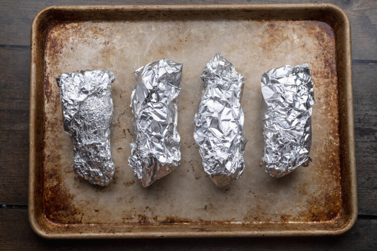 Overhead view of 4 small sweet potatoes wrapped in aluminum foil and lined up horizontally across the center of a rectangular baking sheet.