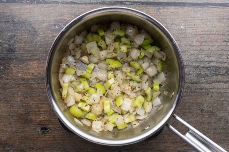 Overhead view of a small silver saucepan containing melted butter, diced onion, diced celery, and seasonings.