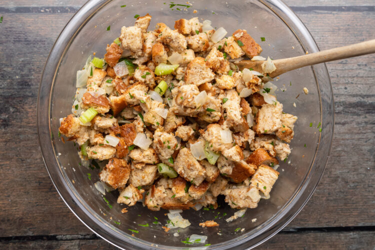 Overhead view of bread stuffing mixture made from cubed bread, seasonings, chicken broth, celery, and onion in a large glass mixing bowl.