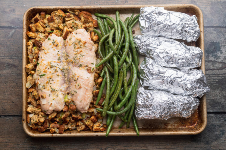 Overhead view of a full baking sheet with turkey, stuffing, green beans, and 4 foil-wrapped sweet potatoes on a wooden countertop.