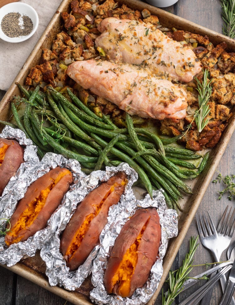 Overhead view of a sheet pan, resting at an angle, filled with green beans, turkey, foil-wrapped sweet potatoes, and stuffing.