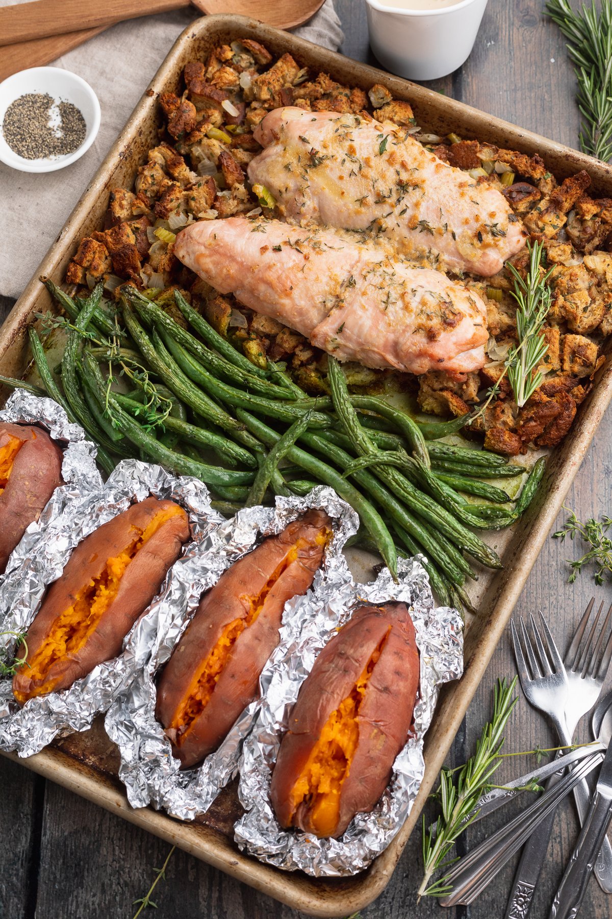 Overhead view of a sheet pan, resting at an angle, filled with green beans, turkey, foil-wrapped sweet potatoes, and stuffing.