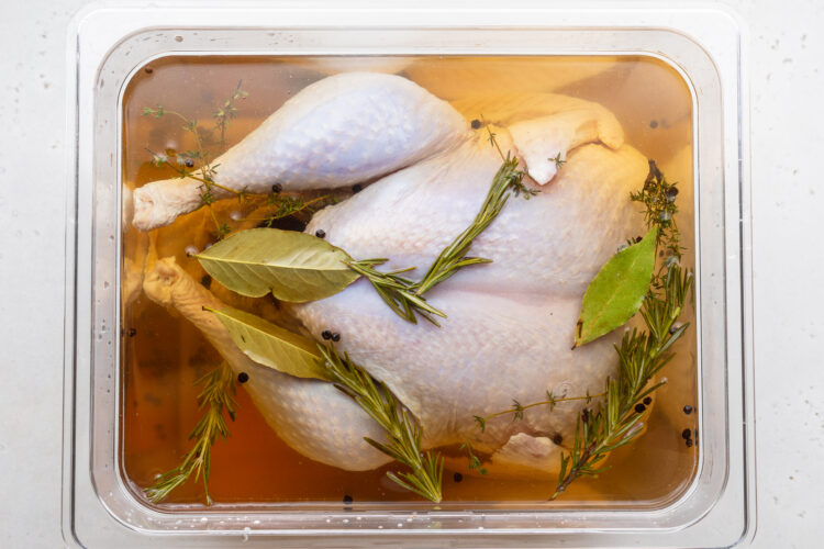 Overhead view of an uncooked turkey in a large rectangular container with a quick turkey brine made from water, brown sugar, and fresh herbs.