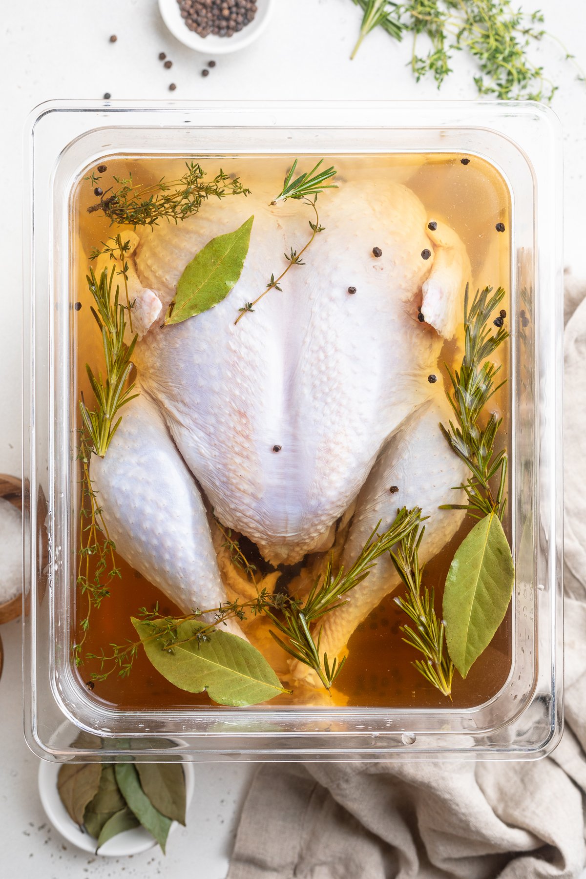 Overhead view of an uncooked whole turkey resting in a container filled with brine and aromatics on a white tabletop.