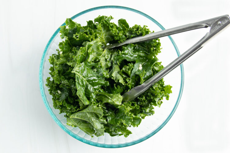 Overhead view of a glass mixing bowl holding large pieces of roughly chopped kale leaves seasoned with oil and garlic.