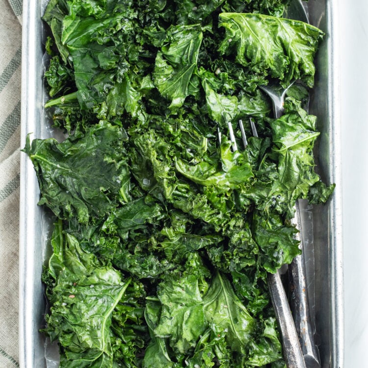 Overhead view of a large sheet pan holding leaves of bright green roasted kale on a neutral tablecloth.