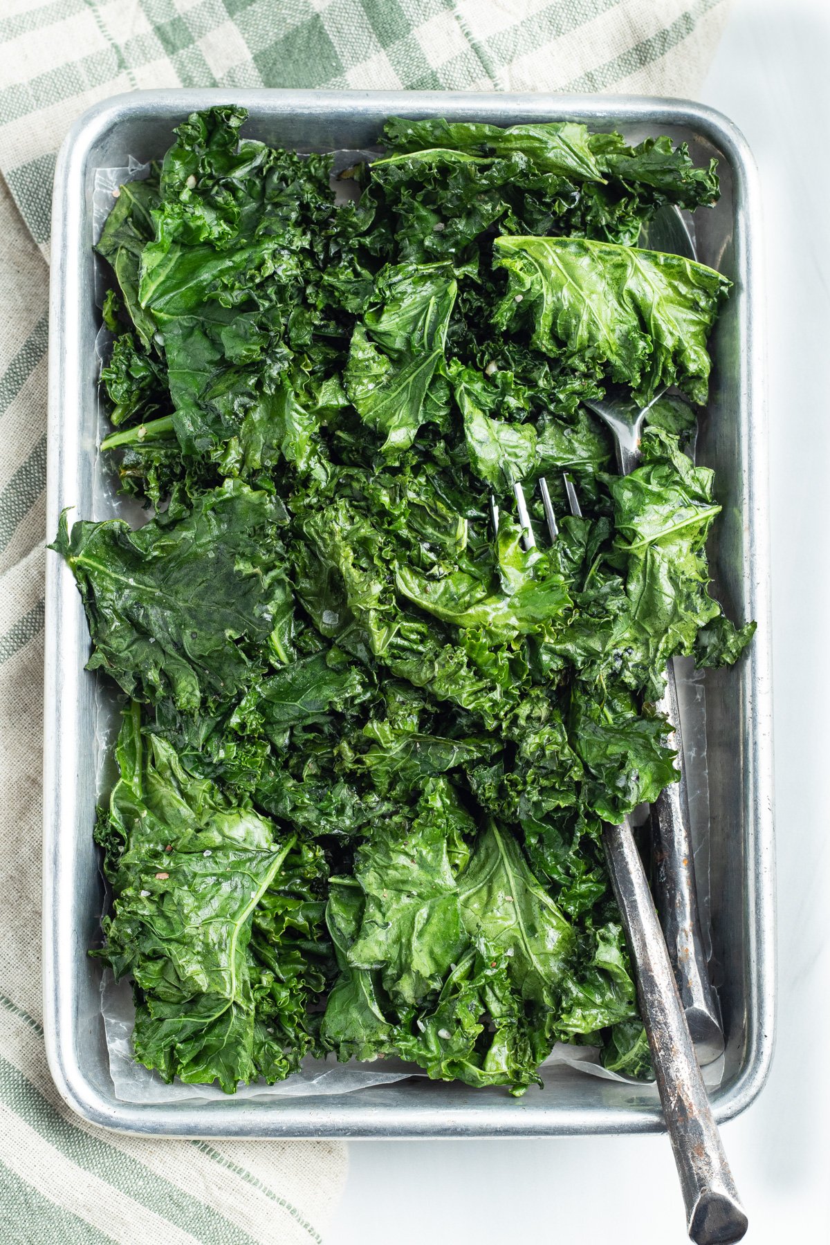 Overhead view of a large sheet pan holding leaves of bright green roasted kale on a neutral tablecloth.