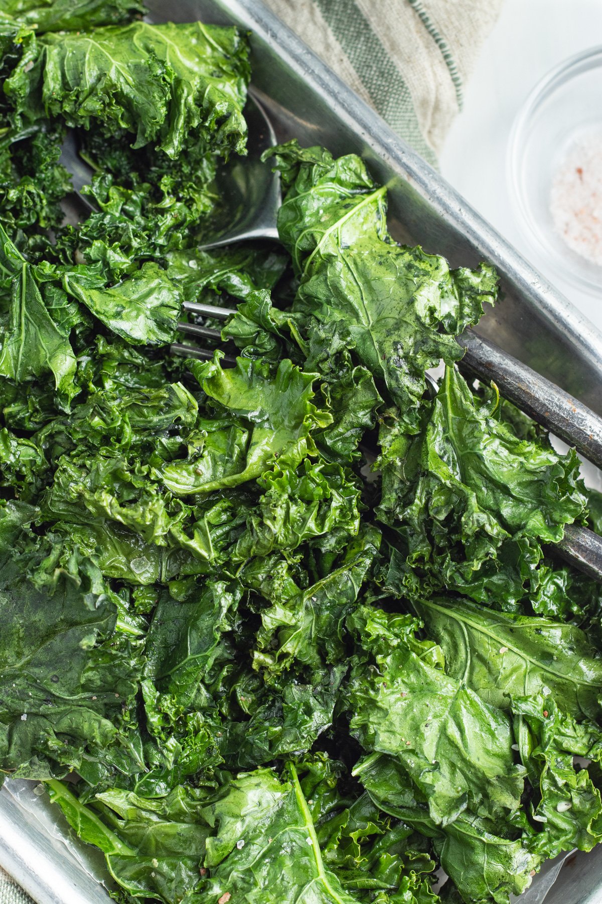 A sheet pan, angled across the field of view, holding bright green leaves of roasted kale with a pair of silver tongs.