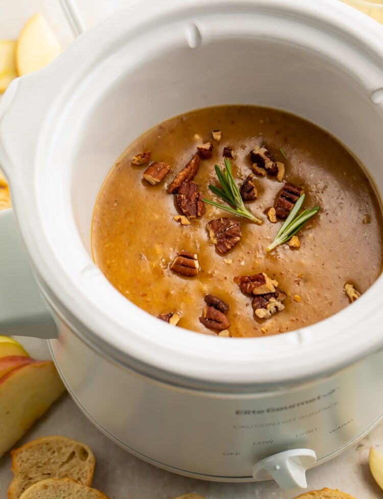 Overhead, angled view of a small white Crockpot slow cooker containing a rich, deep orange baked brie dip topped with pecans and figs.