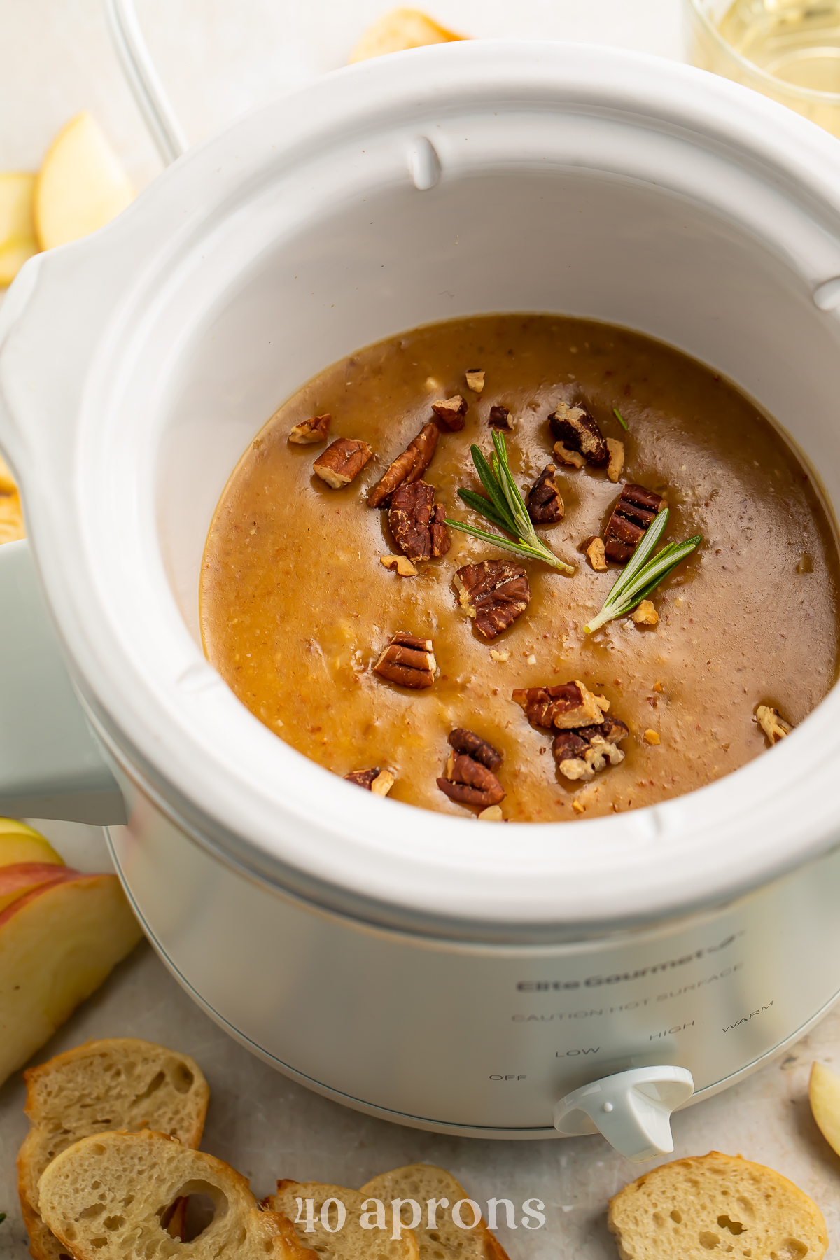 Overhead, angled view of a small white Crockpot slow cooker containing a rich, deep orange baked brie dip topped with pecans and figs.