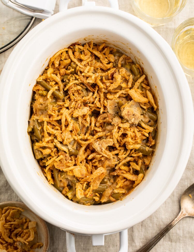 Overhead view of a white oval Crockpot containing a green bean casserole topped with crispy fried onions on a neutral tabletop.