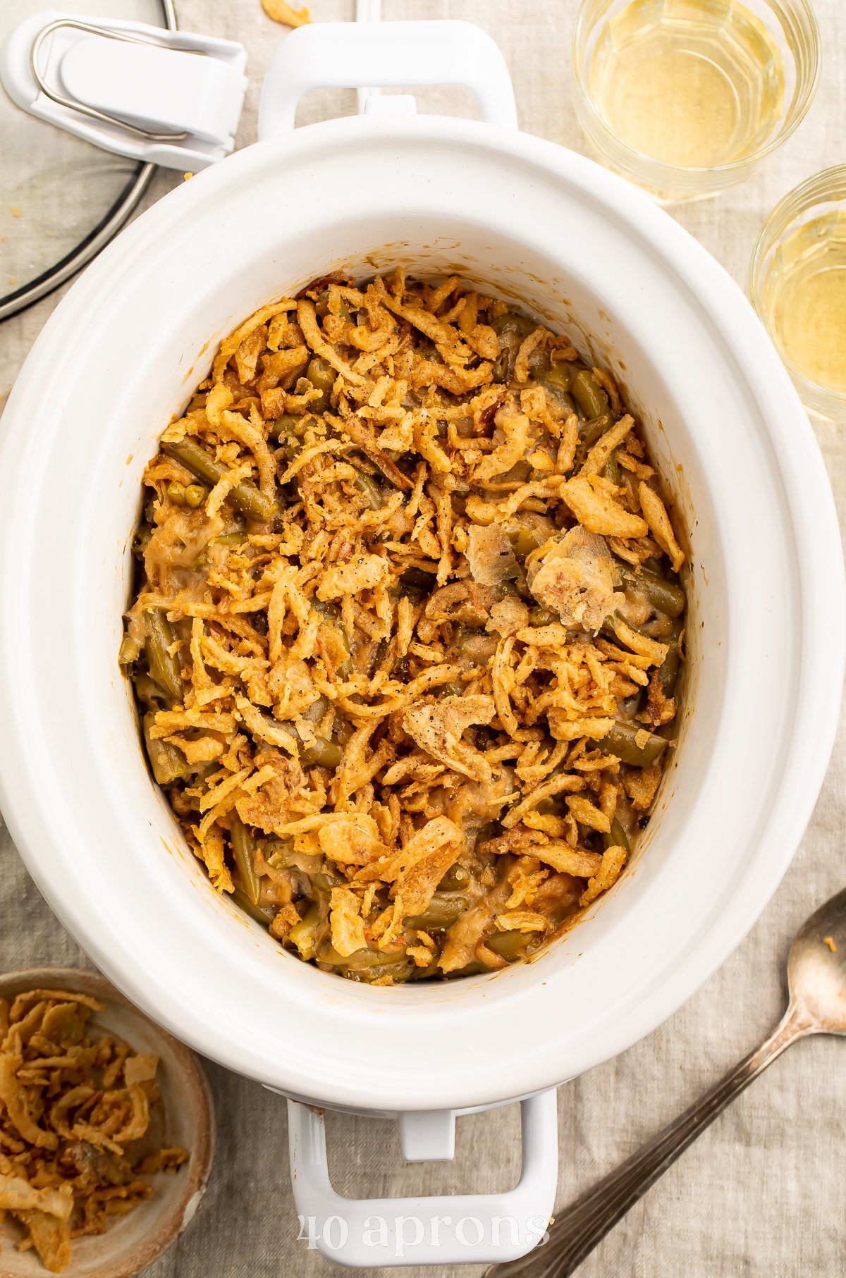 Overhead view of a white oval Crockpot containing a green bean casserole topped with crispy fried onions on a neutral tabletop.