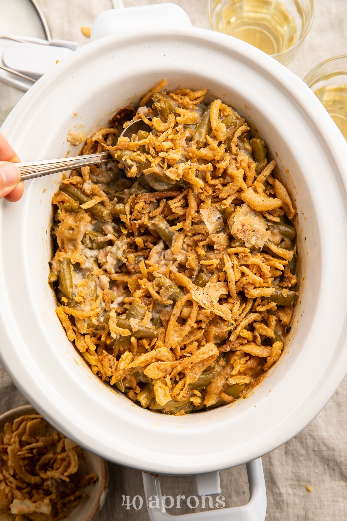 Overhead view of a woman's hand lifting a spoonful of casserole out of a white oval Crockpot containing a green bean casserole topped with crispy fried onions on a neutral tabletop.