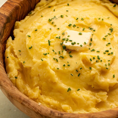 Side-on photo of a wooden bowl of vegan mashed potatoes topped with a square of vegan butter and chopped chives.