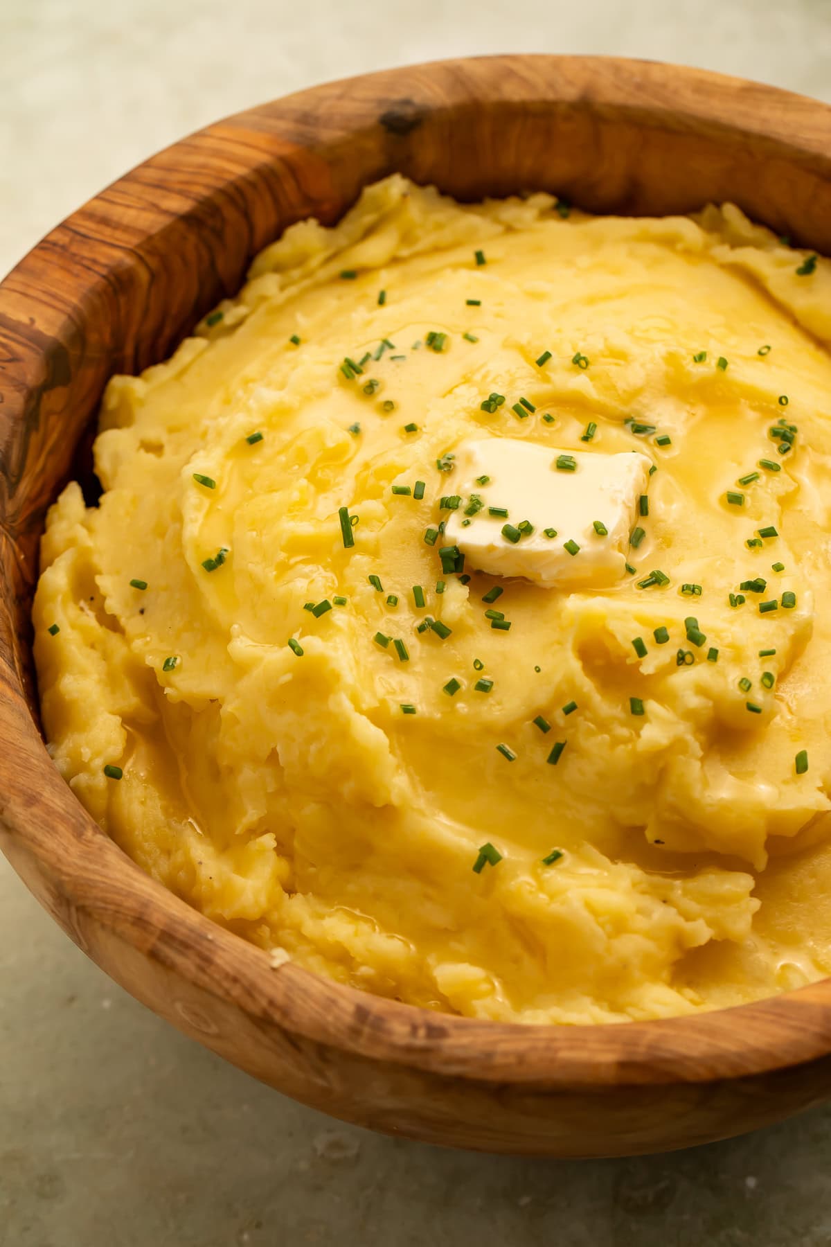 Side-on photo of a wooden bowl of vegan mashed potatoes topped with a square of vegan butter and chopped chives.