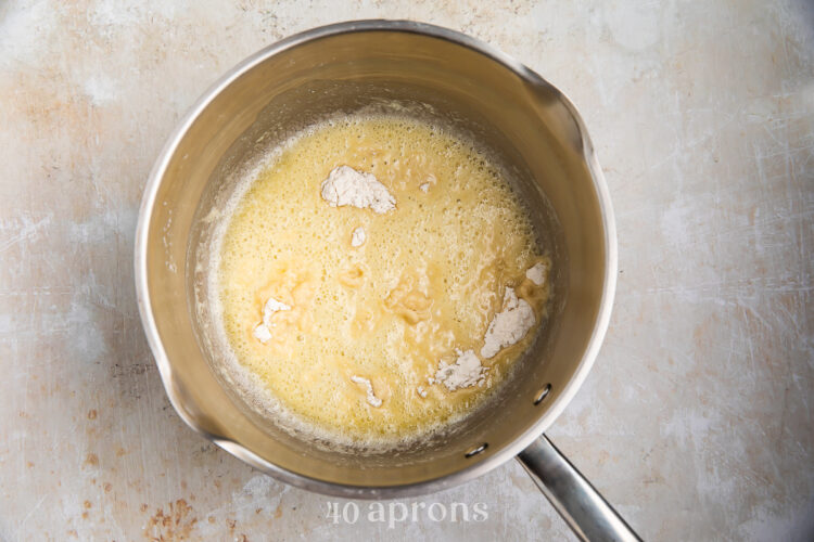 Overhead view of butter and flour for gluten-free gravy in a medium silver saucepan with a handle on a neutral background.