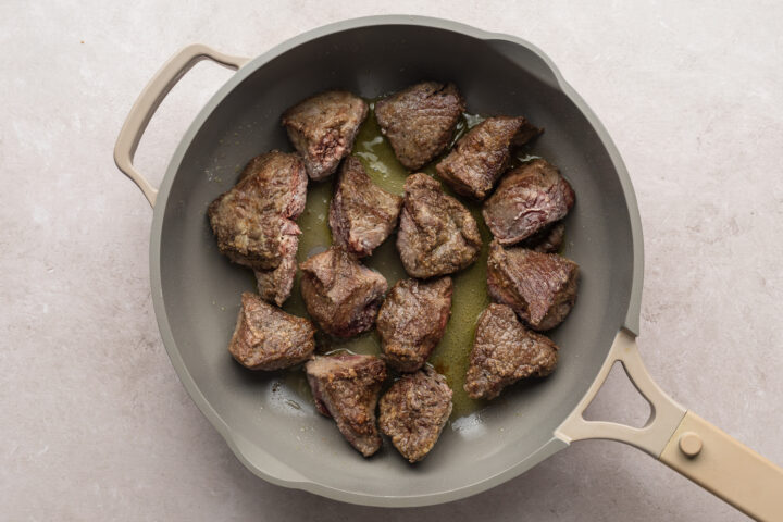 Overhead view of beef tips cooking in a large skillet on a neutral background.