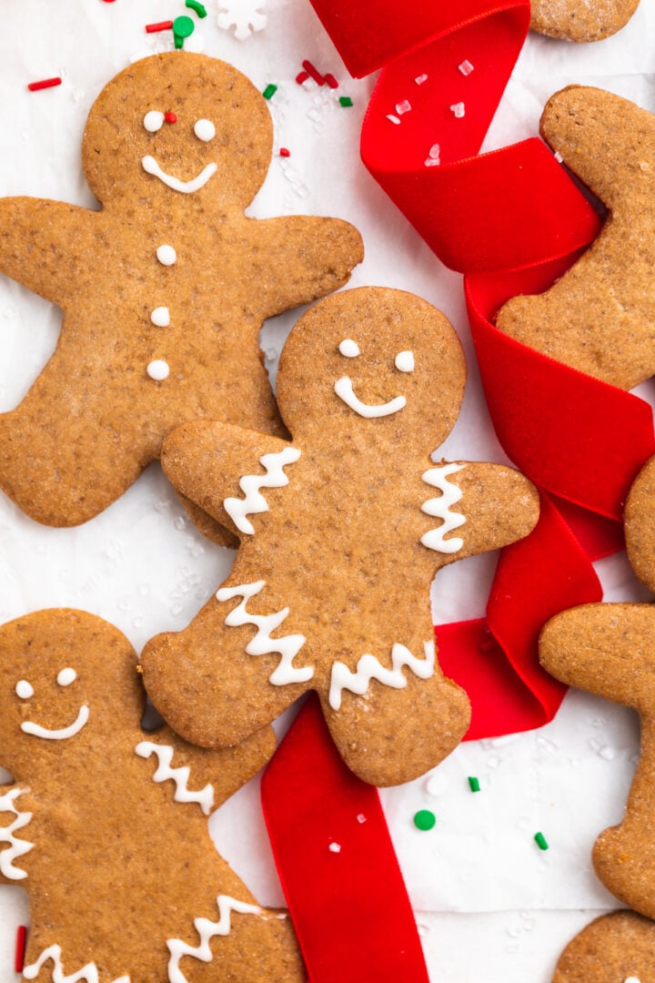 3 gluten-free gingerbread man cookies, decorated with white cooking icing, on a table with red holiday ribbon and sprinkles.