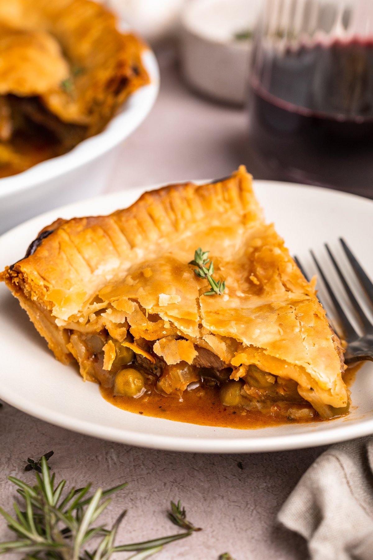 A wedge-shaped slice of beef pot pie on a white plate with a silver fork. The edge of the remaining pot pie is in the background.