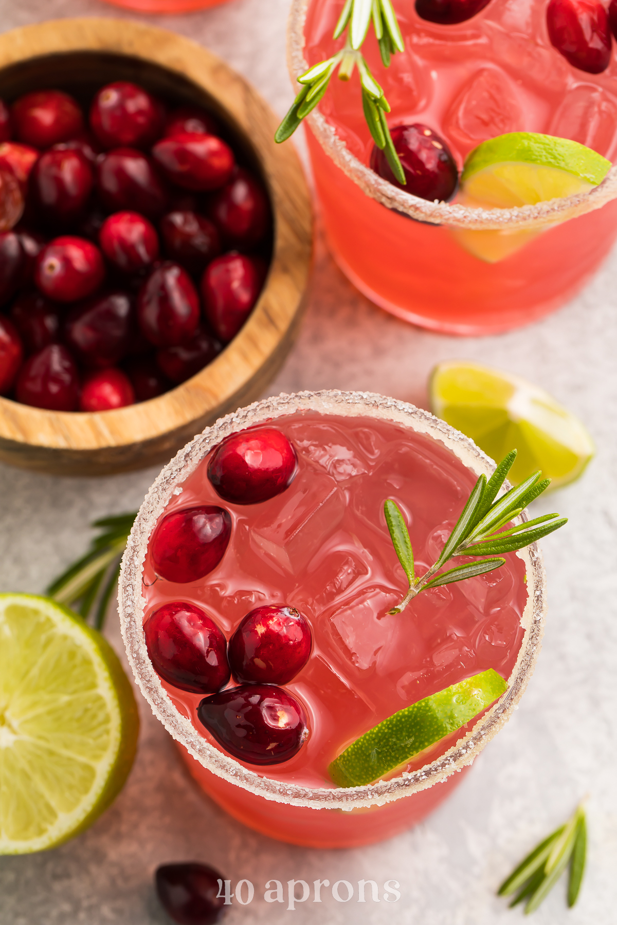 Overhead view of cranberry margaritas on a table. Drinks are bright red and garnished with green rosemary and limes.