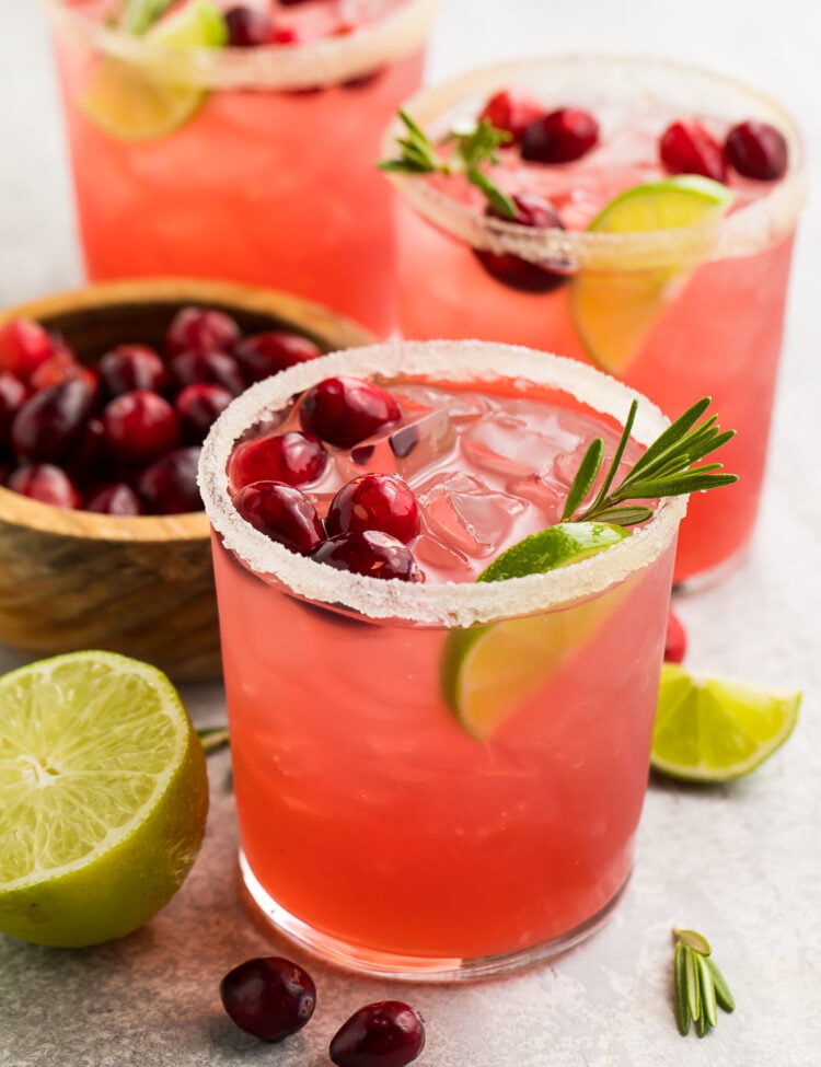 Bright red cranberry margaritas shown in stemless glasses rimmed with salt. Drinks are garnished with cranberries, rosemary, and lime wedges.