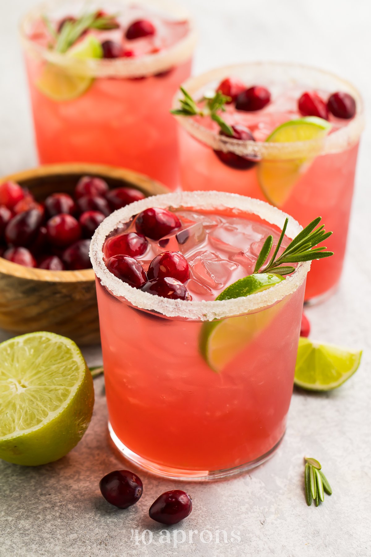 Bright red cranberry margaritas shown in stemless glasses rimmed with salt. Drinks are garnished with cranberries, rosemary, and lime wedges.