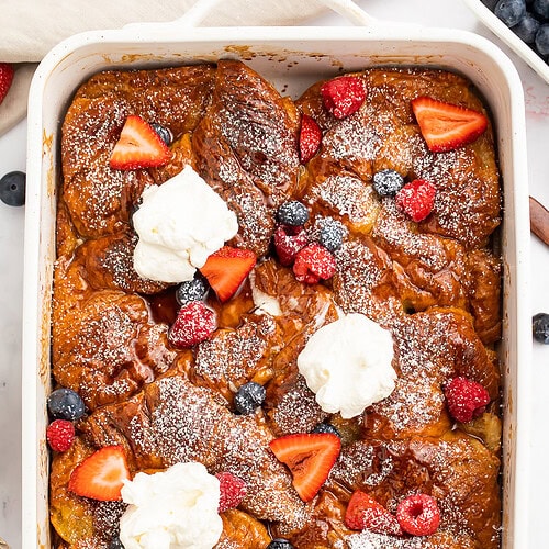 A large casserole dish holding a gorgeous French toast croissant casserole topped with powdered sugar, fresh fruit, and whipped cream.