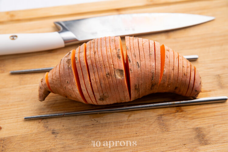 Overhead view of a thinly sliced sweet potato on a wooden cutting board with two skewers on either side and a sharp knife resting above.