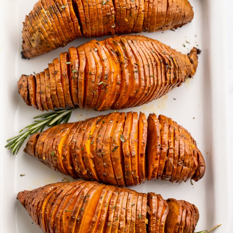 Overhead view of 4 hasselback sweet potatoes lined up in a row vertically down a white rectangular serving board.