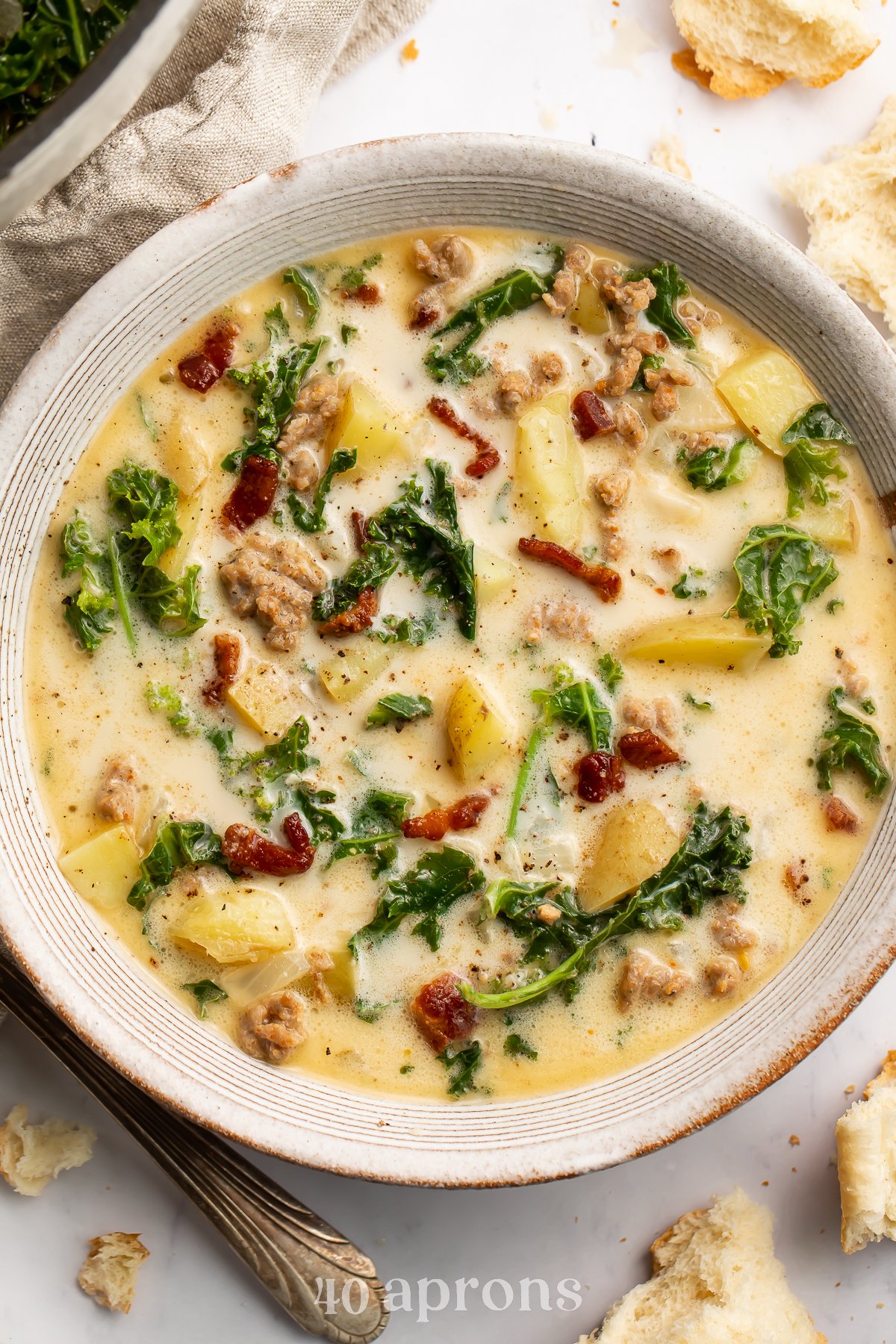 Overhead view of a white soup bowl holding Zuppa Toscana. Floating in the pale yellow soup are spinach, chopped bacon, and chunks of potatoes.