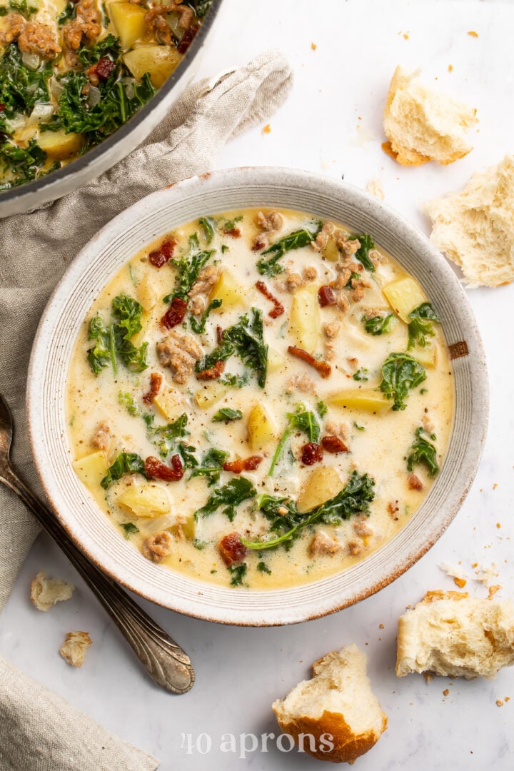 Overhead view of a white soup bowl holding Zuppa Toscana. Floating in the pale yellow soup are spinach, chopped bacon, and chunks of potatoes.