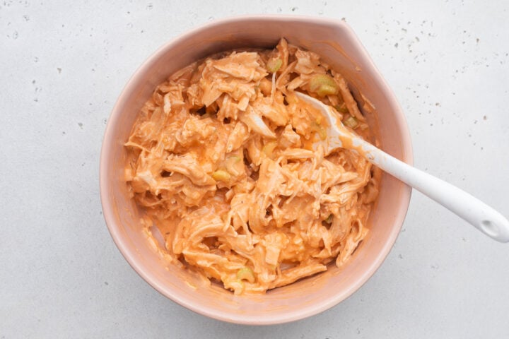 Ingredients for Whole30 buffalo chicken salad mixed together in a large white mixing bowl on a white background.