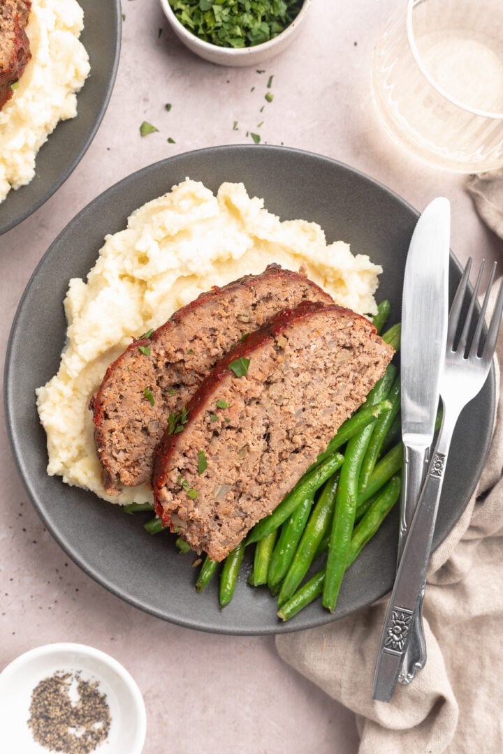 Two slices of Whole30 meatloaf plated with green beans and mashed potatoes on a grey-blue plate with silverware on a table.