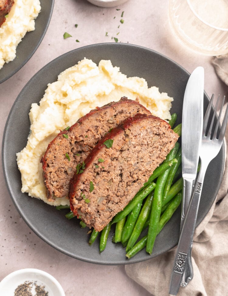 Two slices of Whole30 meatloaf plated with green beans and mashed potatoes on a grey-blue plate with silverware on a table.