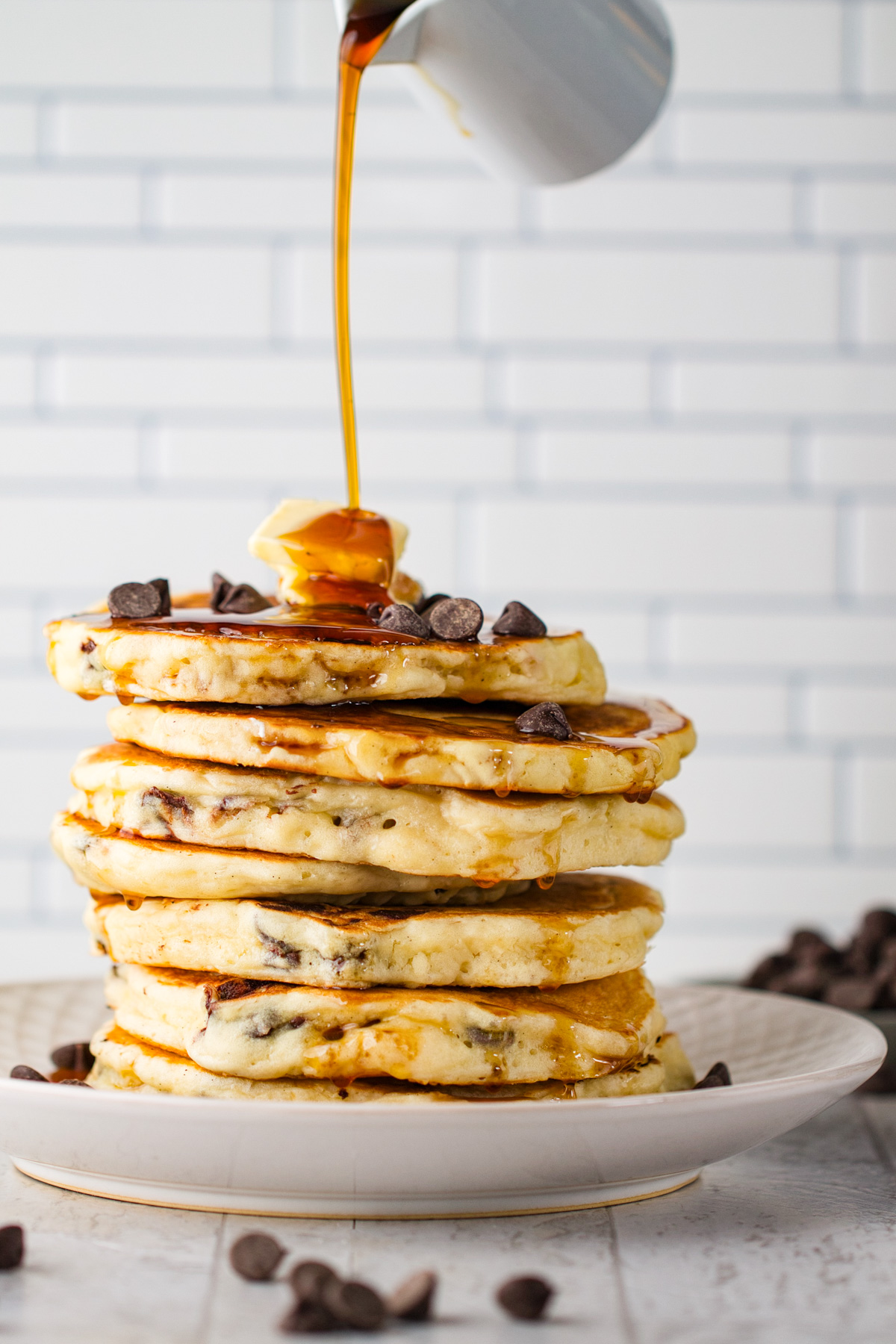 Syrup being poured over a stack of chocolate chip pancakes topped with pats of butter.