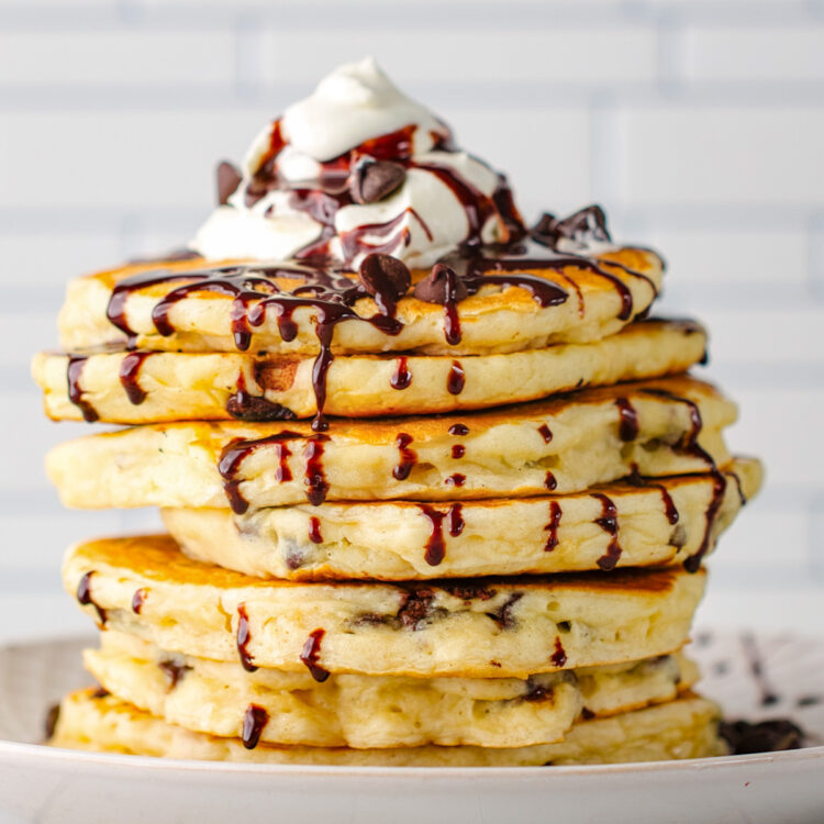 A stock of chocolate chip pancakes topped with whipped cream, with chocolate sauce drizzled over the top and down the sides of the pancakes.
